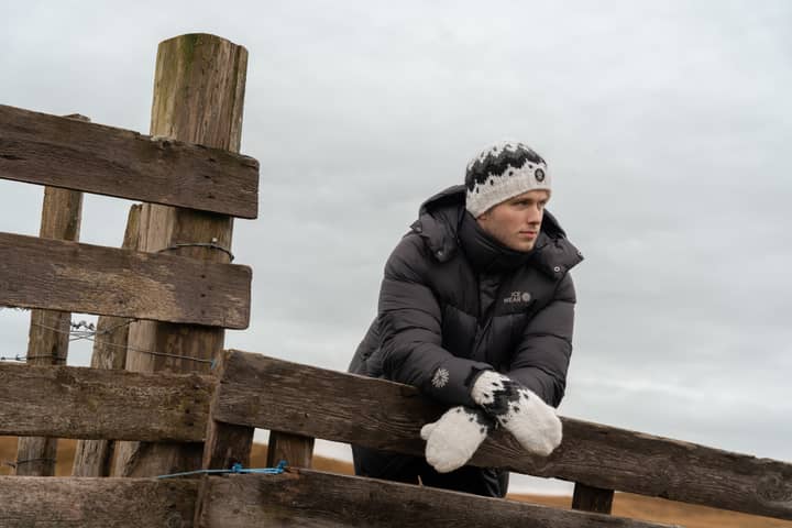 A man standing by a fence, wearing a warm coat with a hat and mittens