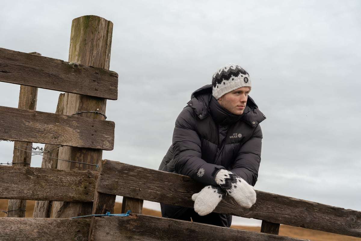 A man standing by a fence, wearing a warm coat with a hat and mittens
